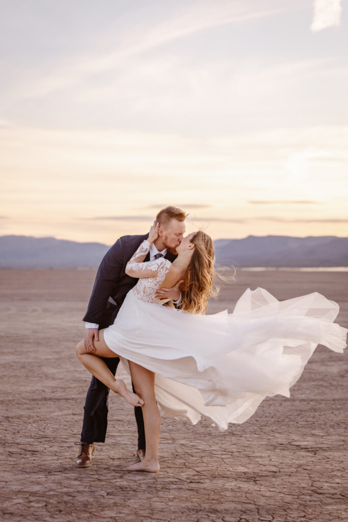 Groom dips bride for a kiss at a dry lake bed in Las Vegas during sunset. Her dress flows beautifully in the wind behind her. 