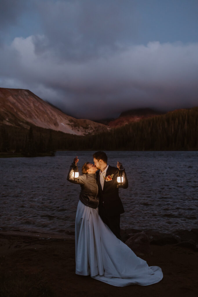 Bride and groom stand next to each other holding lanterns during blue hour as the sun rises at long lake