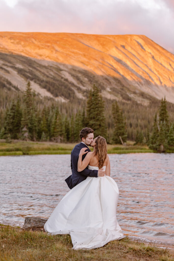 bride and groom kiss while admiring the alpen glow during their adventure session at long lake during sunrise